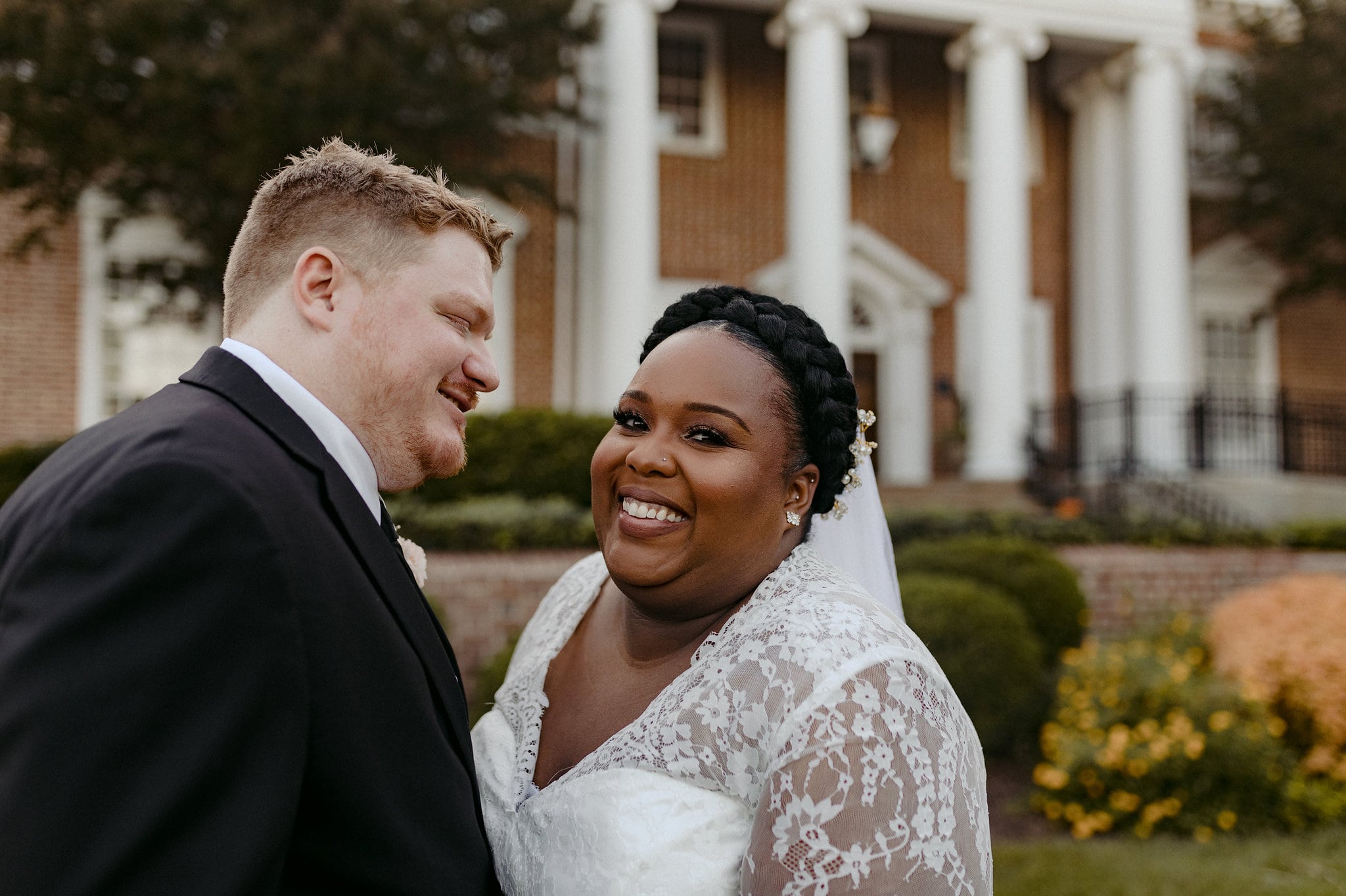a mixed-race couple in wedding clothes smiles in front of a brick building with white columns
