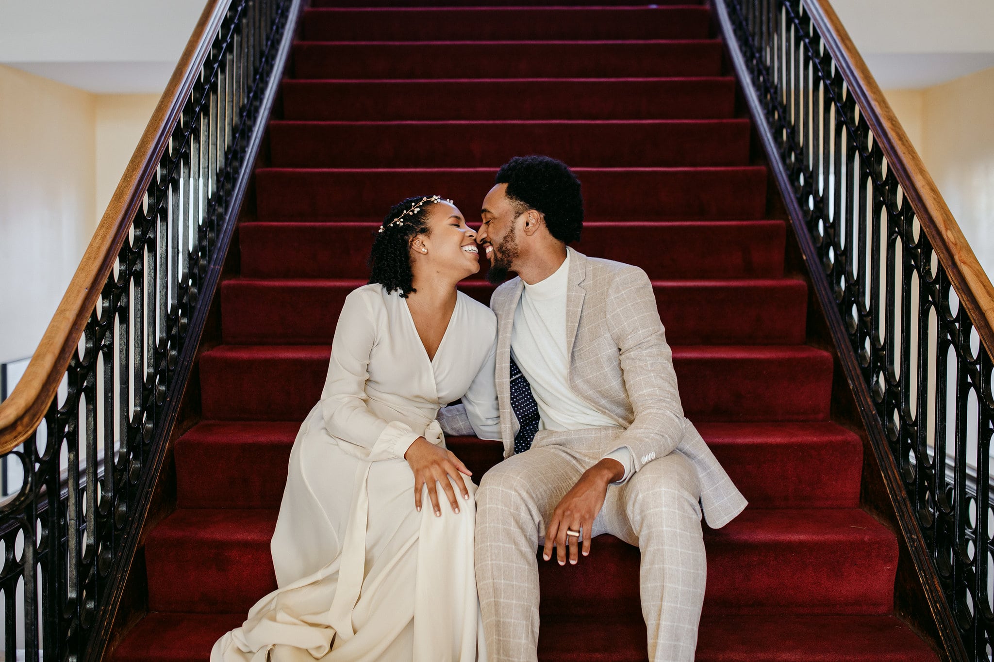 a couple in wedding attire sit on dark red carpeted steps and smile at each other, faces almost touching