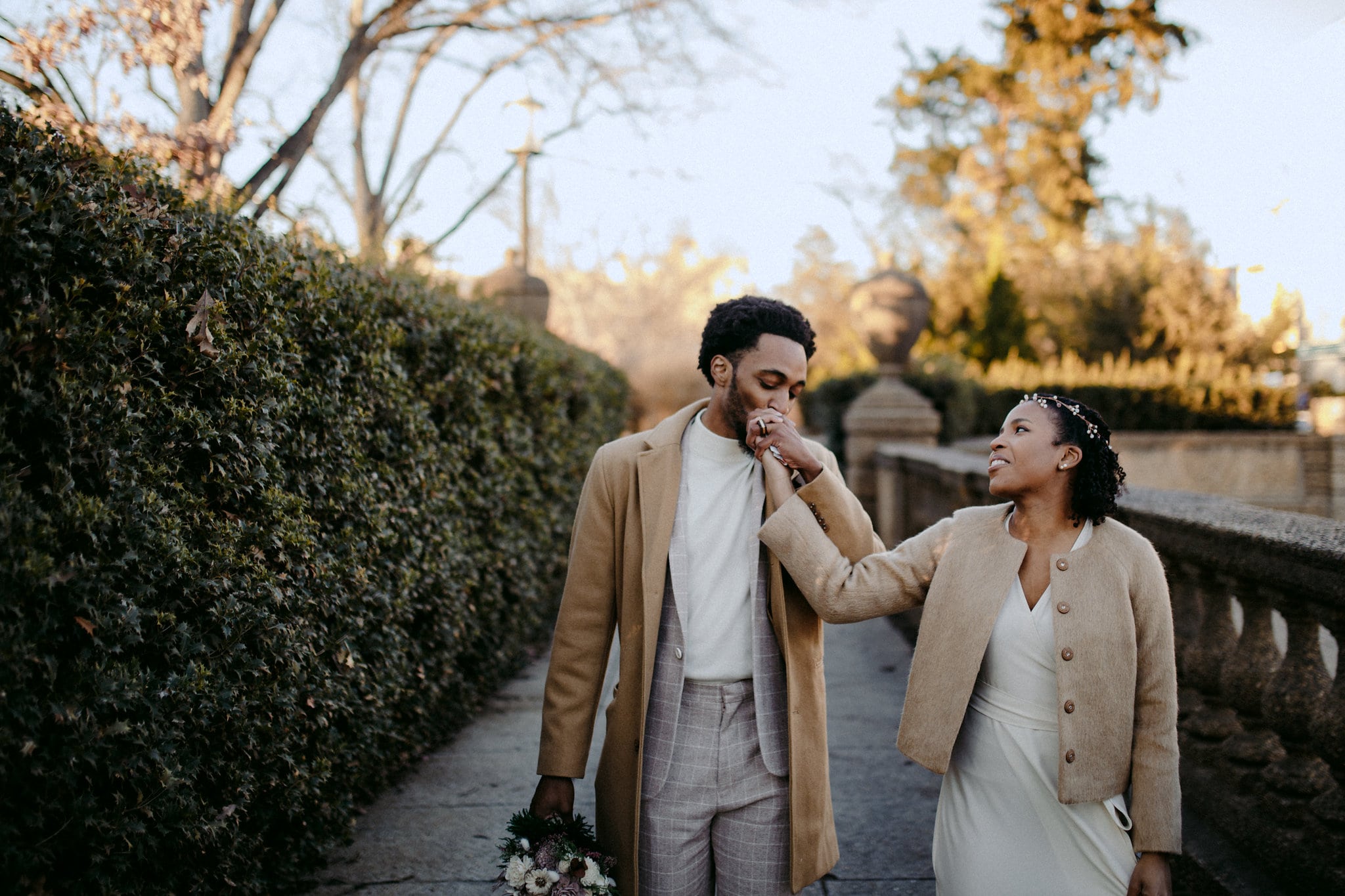 a groom kisses a brides hand and holds her bouquet as they walk in a park wearing jackets on a cold day