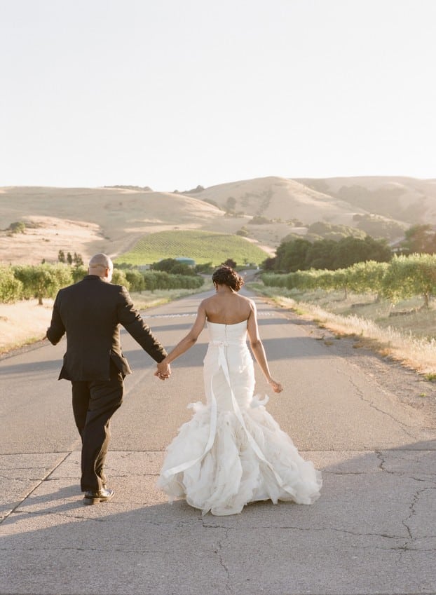A man and a woman in wedding clothes hold hands and walk down a road quiet together in wine country