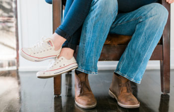 A woman sitting on a man's lap in a wooden arm chair; we only see their legs in jeans and shoes
