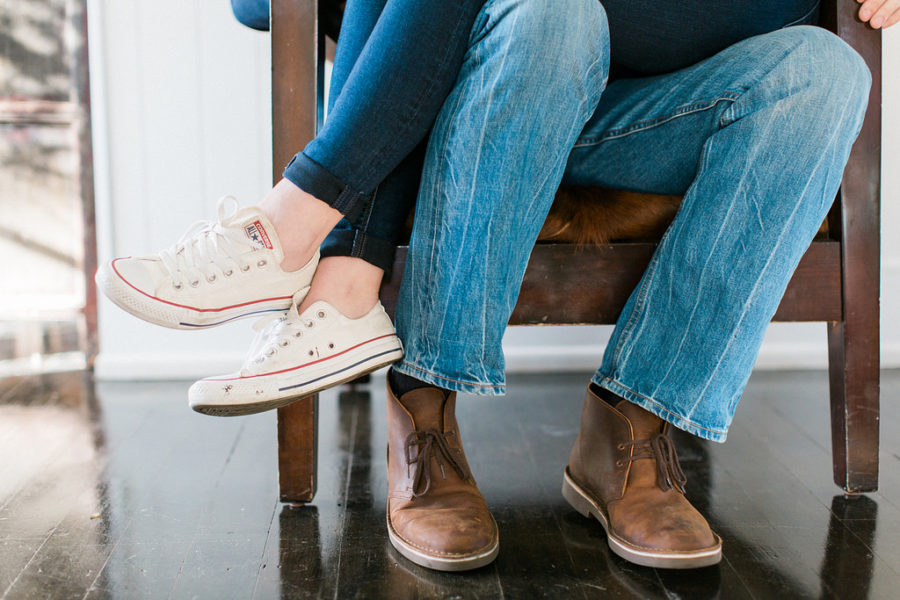 A woman sitting on a man's lap in a wooden arm chair; we only see their legs in jeans and shoes