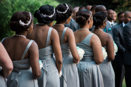 Five African-American bridesmaids with their hair up wearing light grey dresses and holding bouquets standing in a row with their backs to you during a wedding ceremony