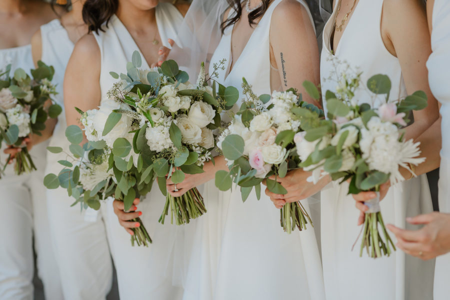 Women in all white sleeveless dresses and jumpsuits, holding bouquets