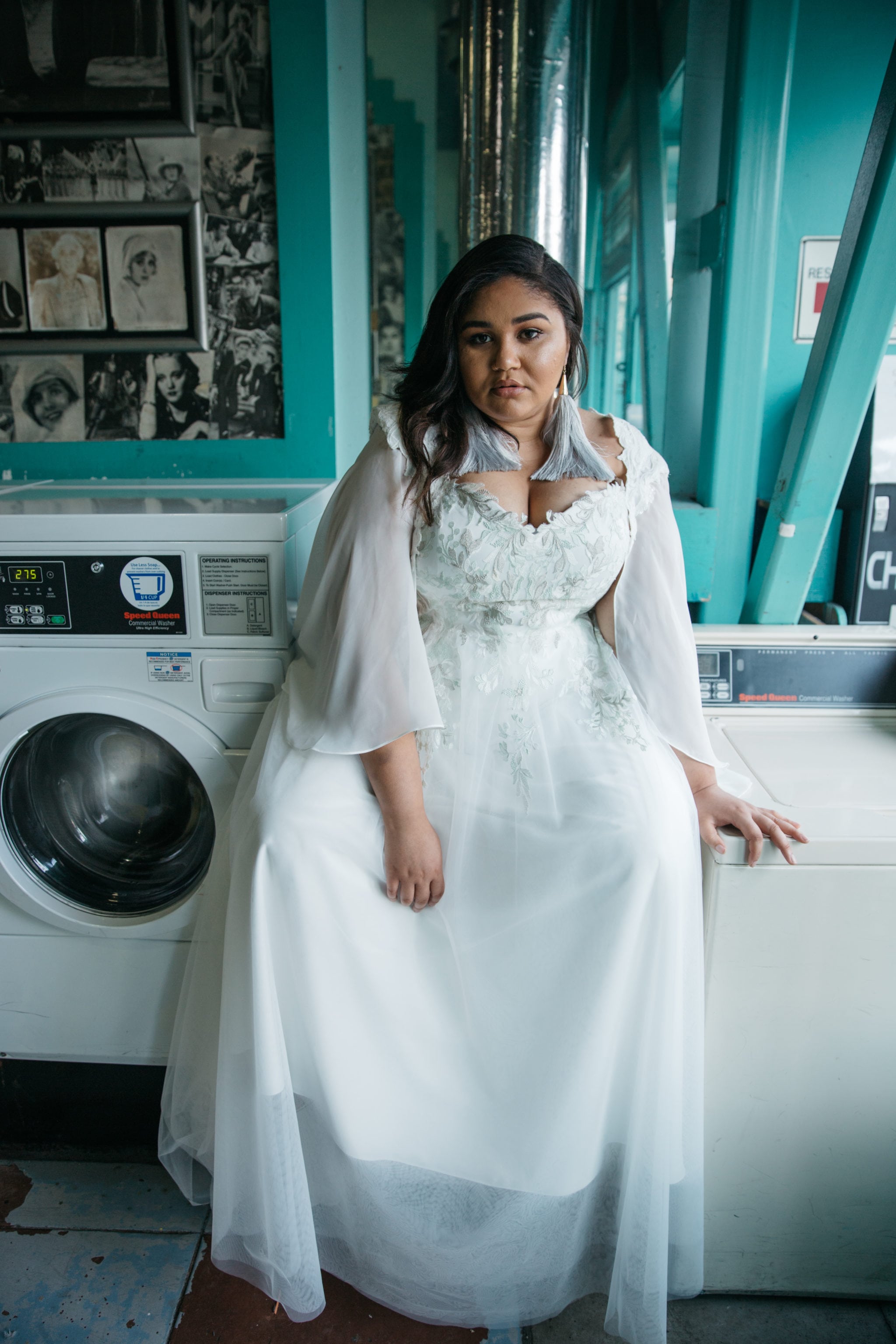 A woman wearing a plus size wedding dress and capelet from Lace and Liberty sits on a washing machine in a laundromat with teal walls