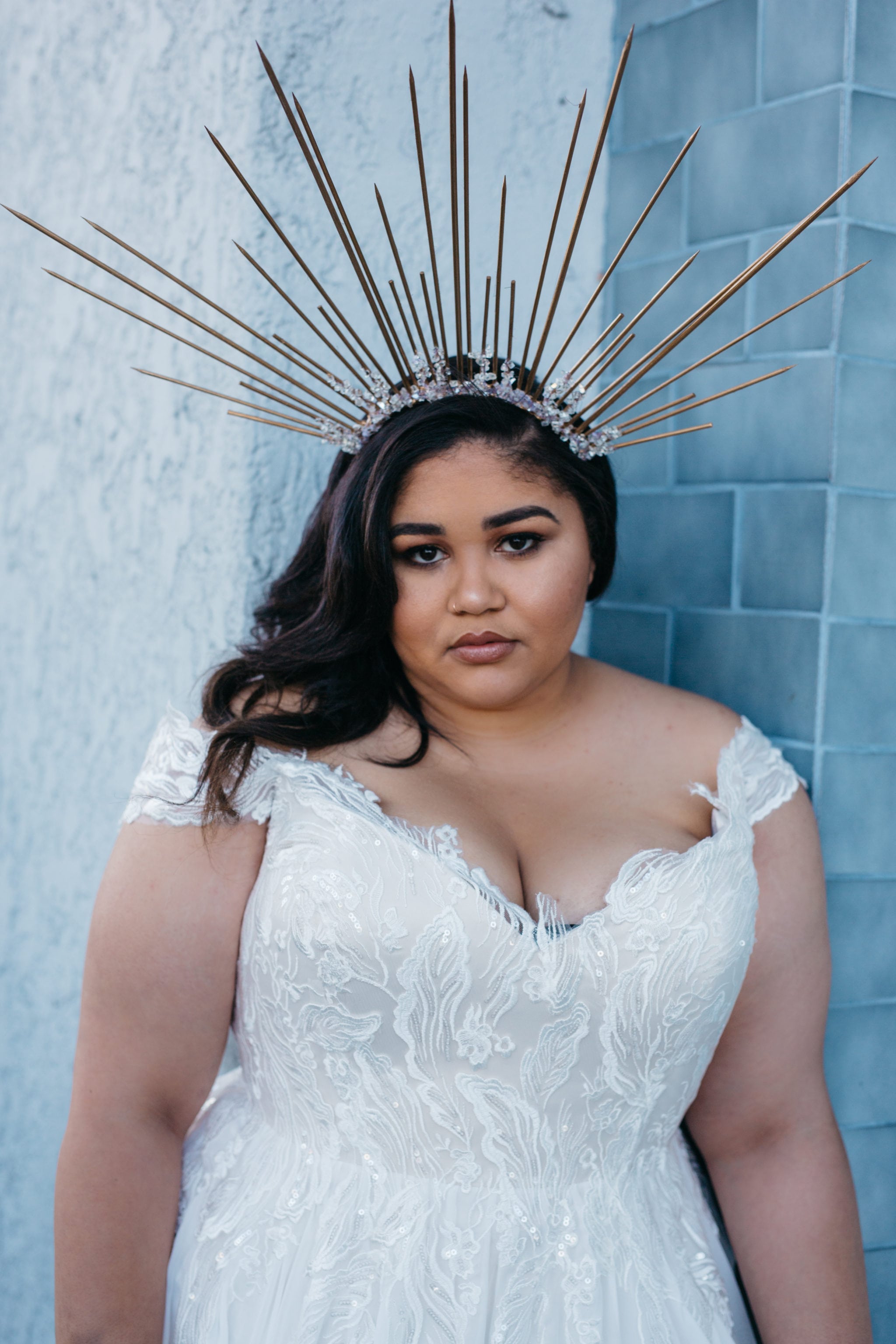 A woman wearing plus size wedding dresses from Lace and Liberty with a lace bodice and deep V neckline and a radiant crown leans against a blue-grey tile wall and looks at you 