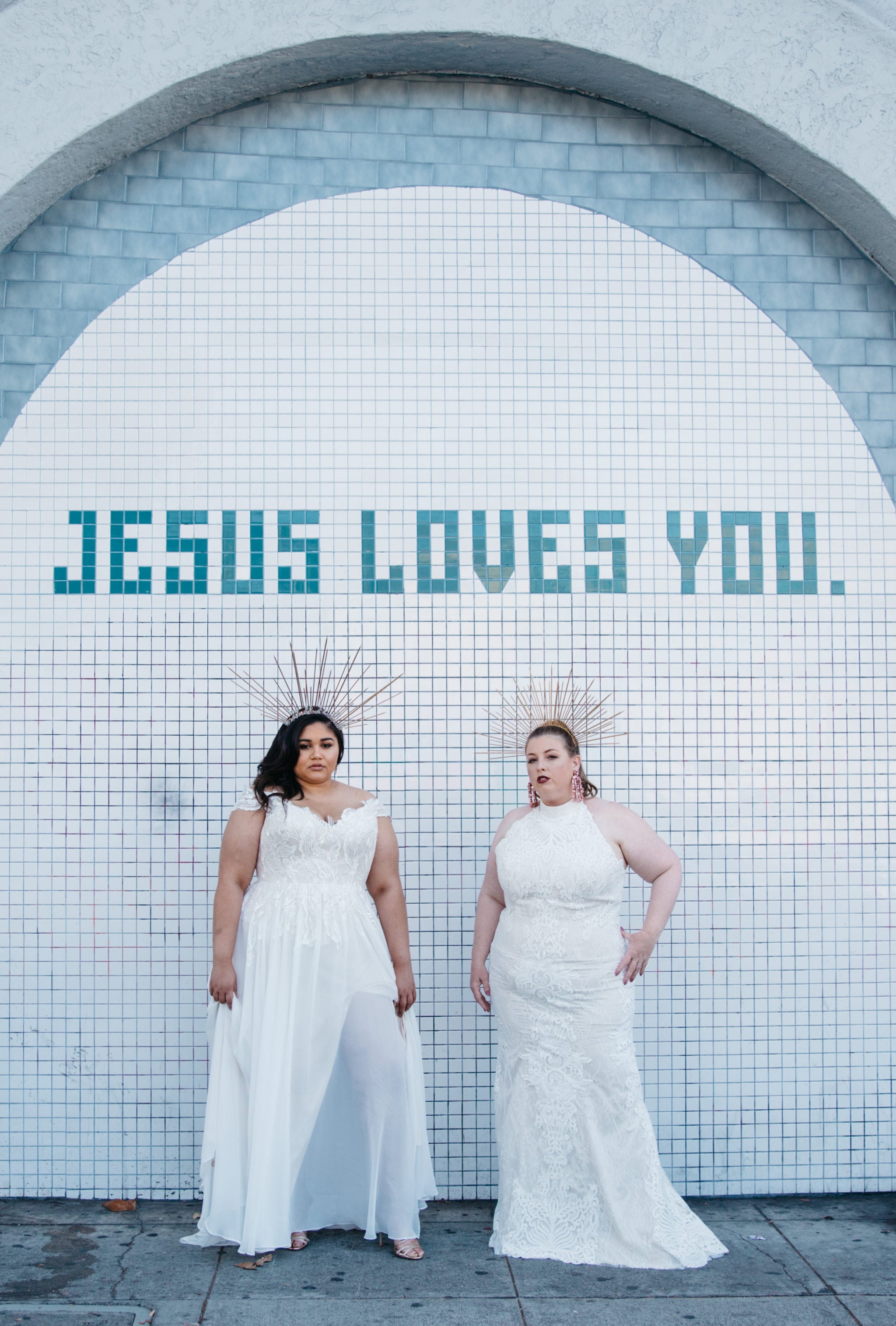 Two women wearing plus size wedding dresses from Lace and Liberty stand in front of a tile wall that reads "Jesus loves you." 