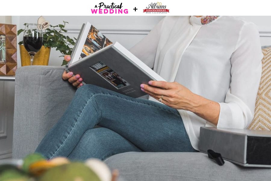 A woman in a white sweater and jeans sits on a couch looking through a grey leather-bound photo album from Albums Remembered