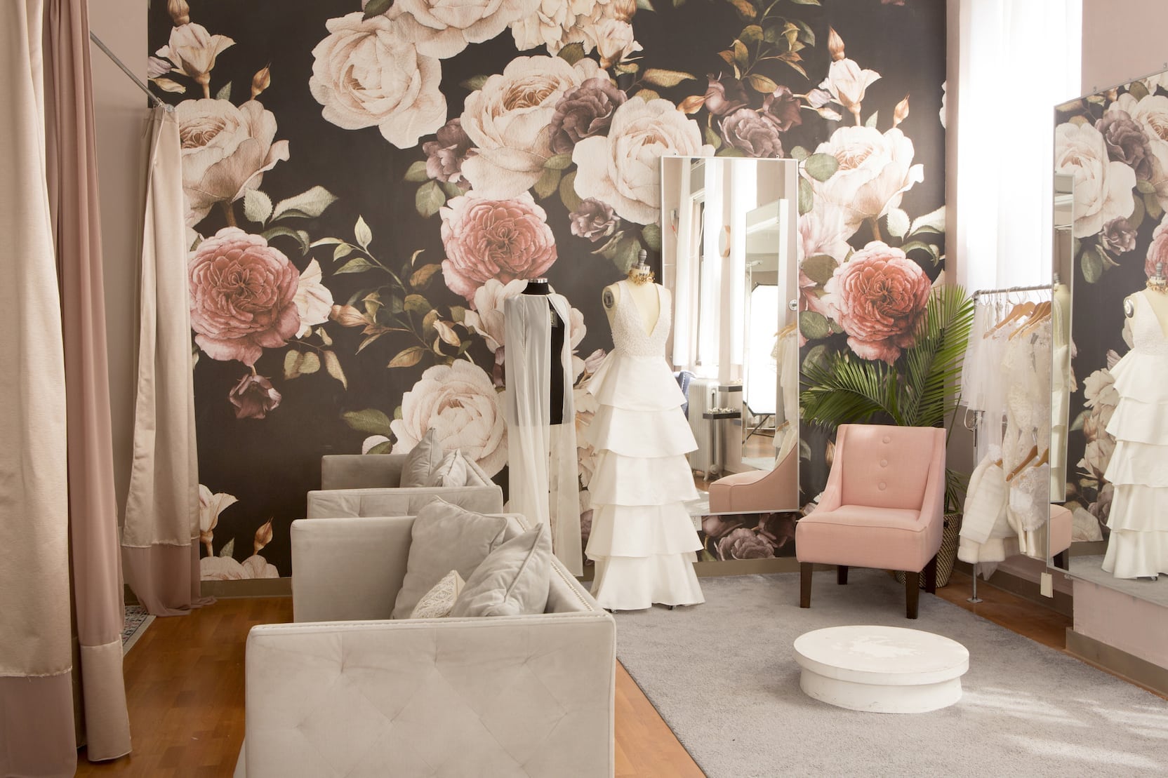 A fitting room with a large floral wall paper, modern grey sofas, and a millennial pink velvet chair.