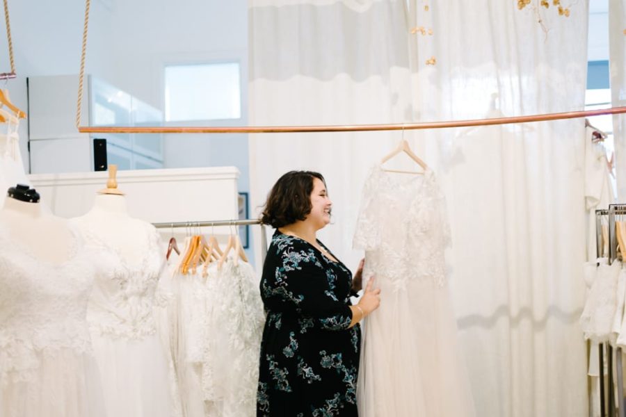 plus size brunette woman wearing a dark floral print dress touching her wedding dress for the first time at Lace and Liberty and smiling