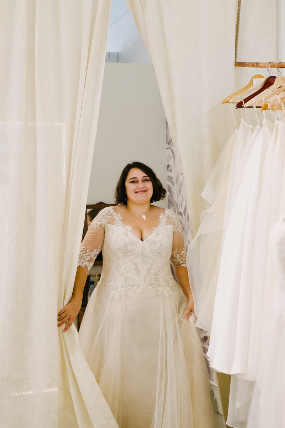 plus size brunette woman walking out of the dressing room with her white, v neck lace wedding dress on and a smile on her face at Lace and Liberty