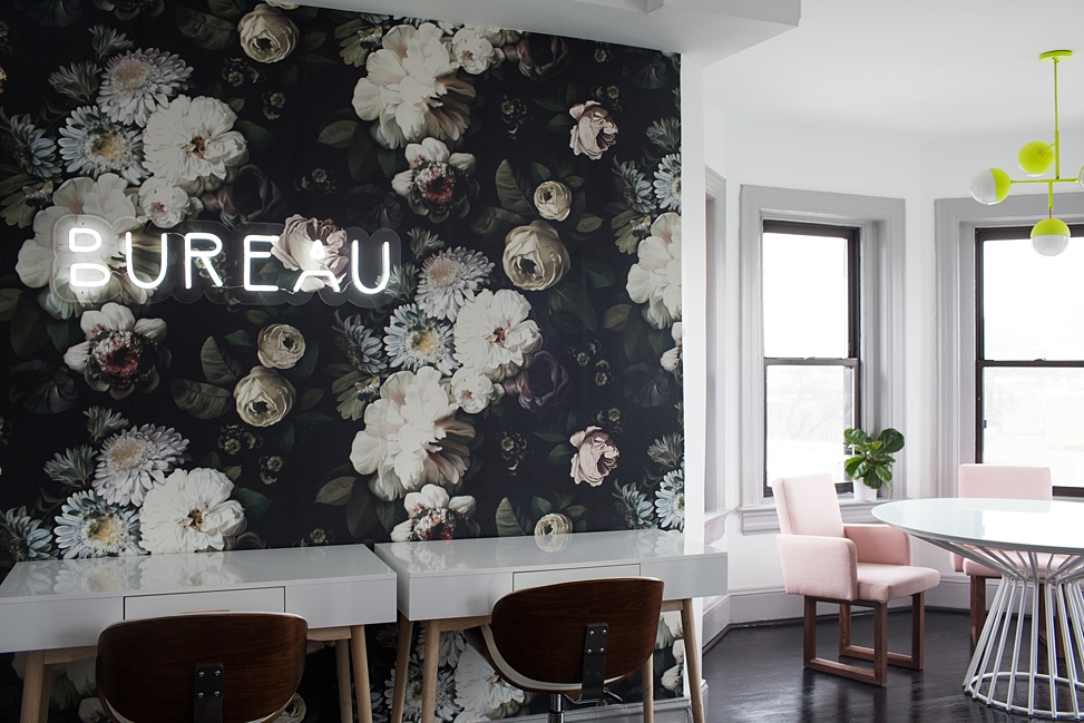 The interior of the Bureau co-working space with a dark floral wall-papered wall with a white neon sign that reads "Bureau," glossy white tables and an alcove with Millennial pink chairs and a white table