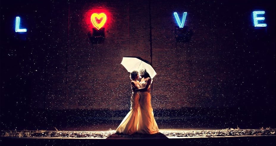 Two brides embrace under an umbrella in the rain beneath a neon sign that reads "Love"