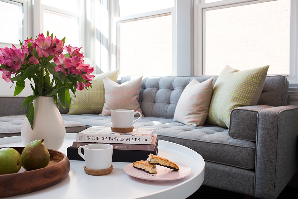Interior of the Bureau co-working lounge area witha grey couch, white round coffee table, vase of pink flowers, tray of pears, two coffees and biscotti