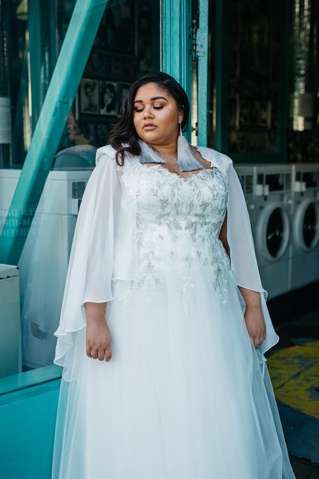 A woman in a plus size wedding dress with an embroidered bodice and capelet from Lace & Liberty stands in the entrance of a laundromat