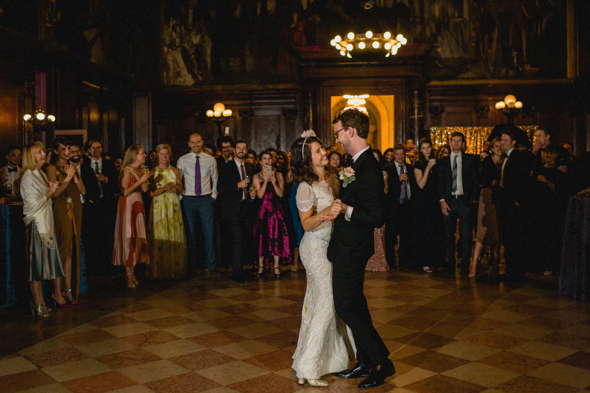 A bride and groom dancing in a dark wood paneled ballroom, surrounded by friends and family