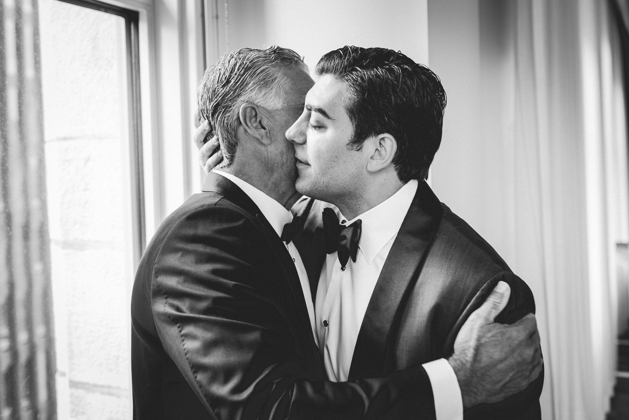 two men in tuxedos and bowties hug tightly in a black and white image