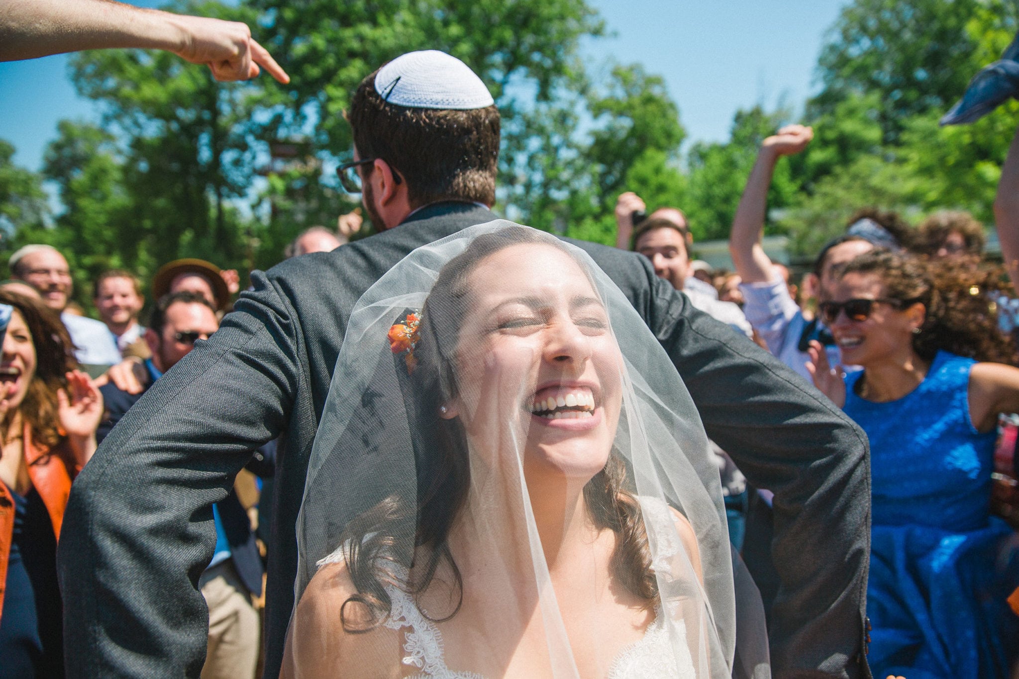 a bride under a veil laughs with her eyes closed with her back to the groom in a yarmulke, surrounded by family and friends outside