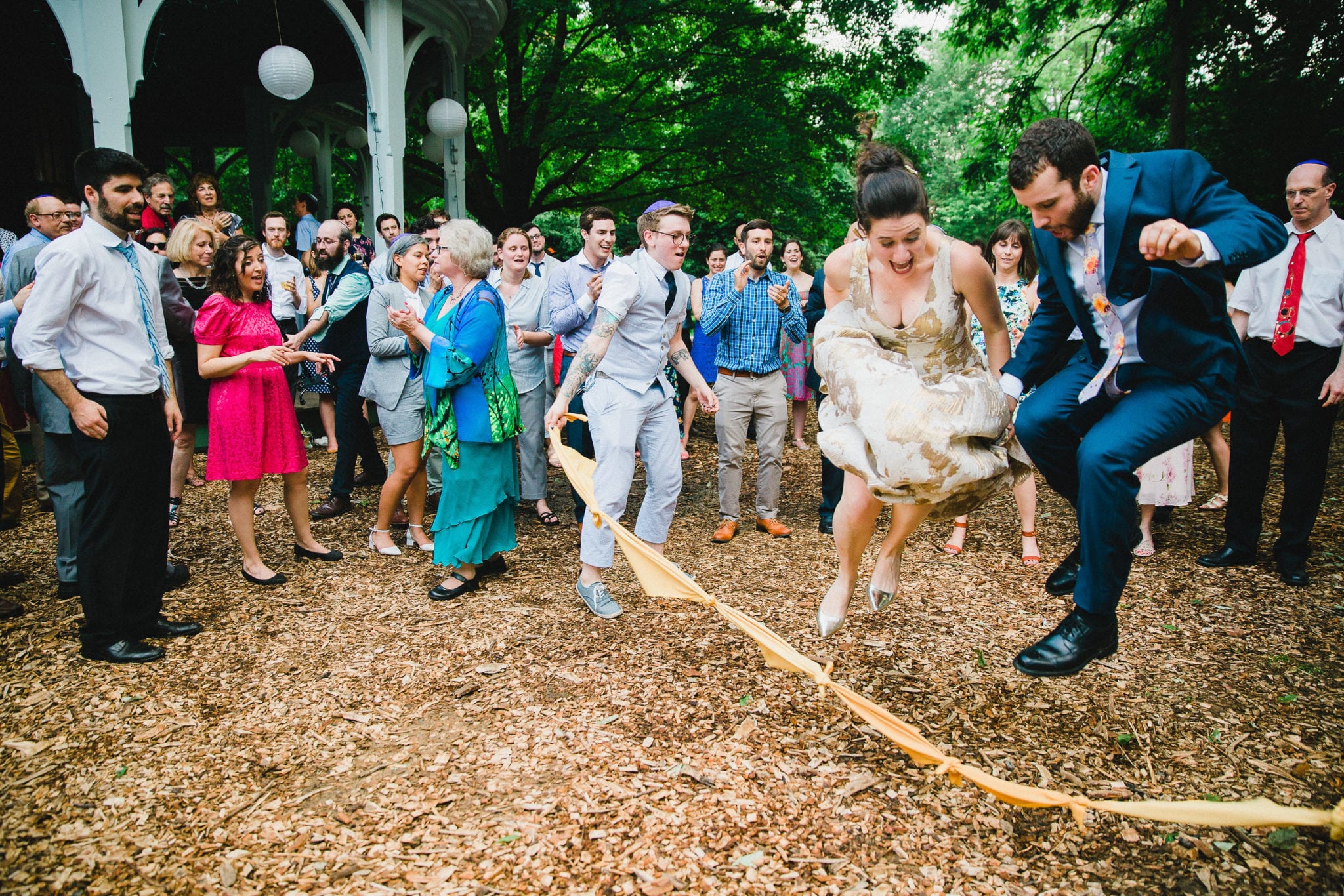 a bride and groom jump rope at an outdoor wedding reception as friends and family look on