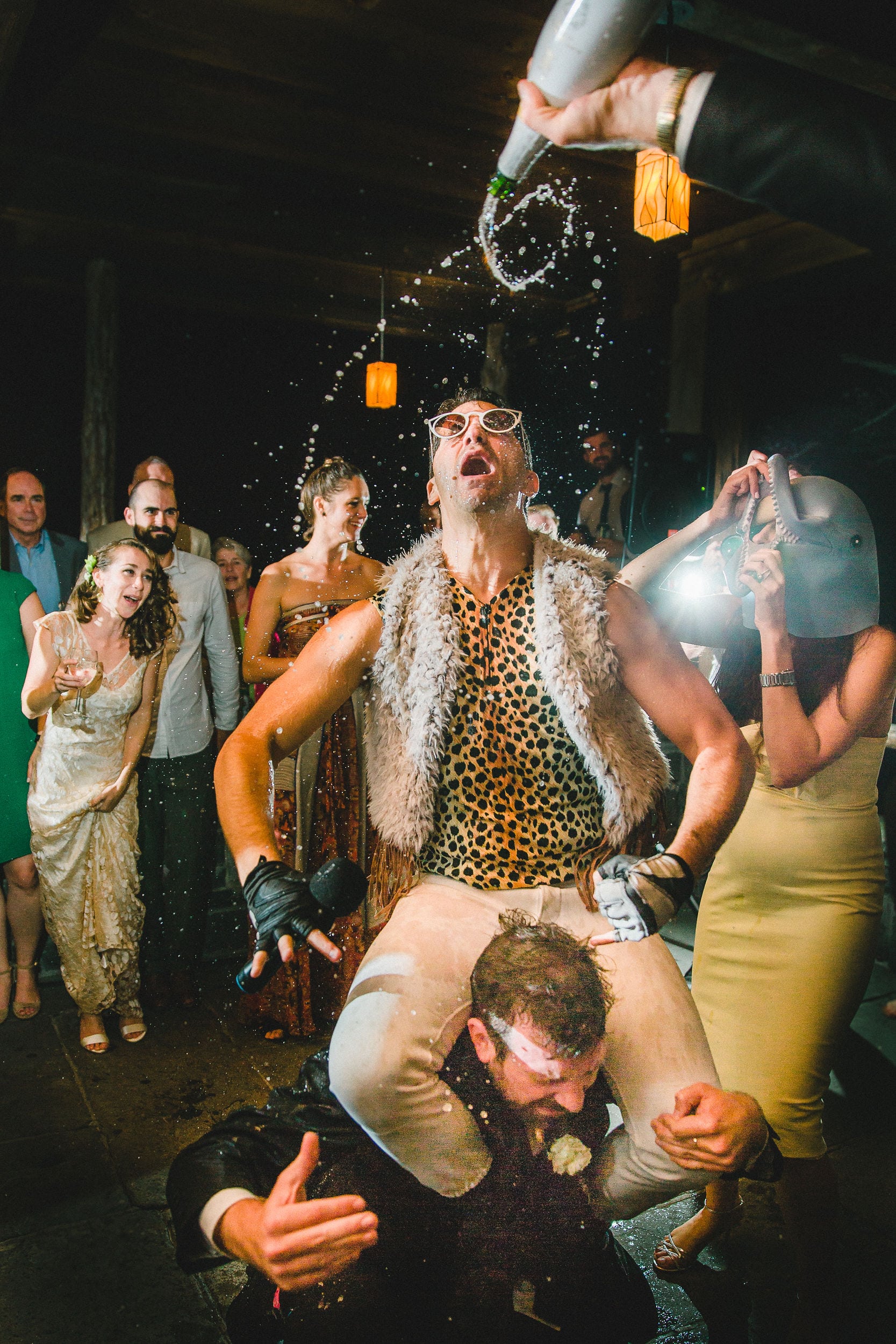 a man in a leopard print sleeveless shirt, fur vest, sunglasses, and black leather fingerless gloves is doused with a bottle of champagne as he sits on a man in a tux's shoulders at an outdoor party at night