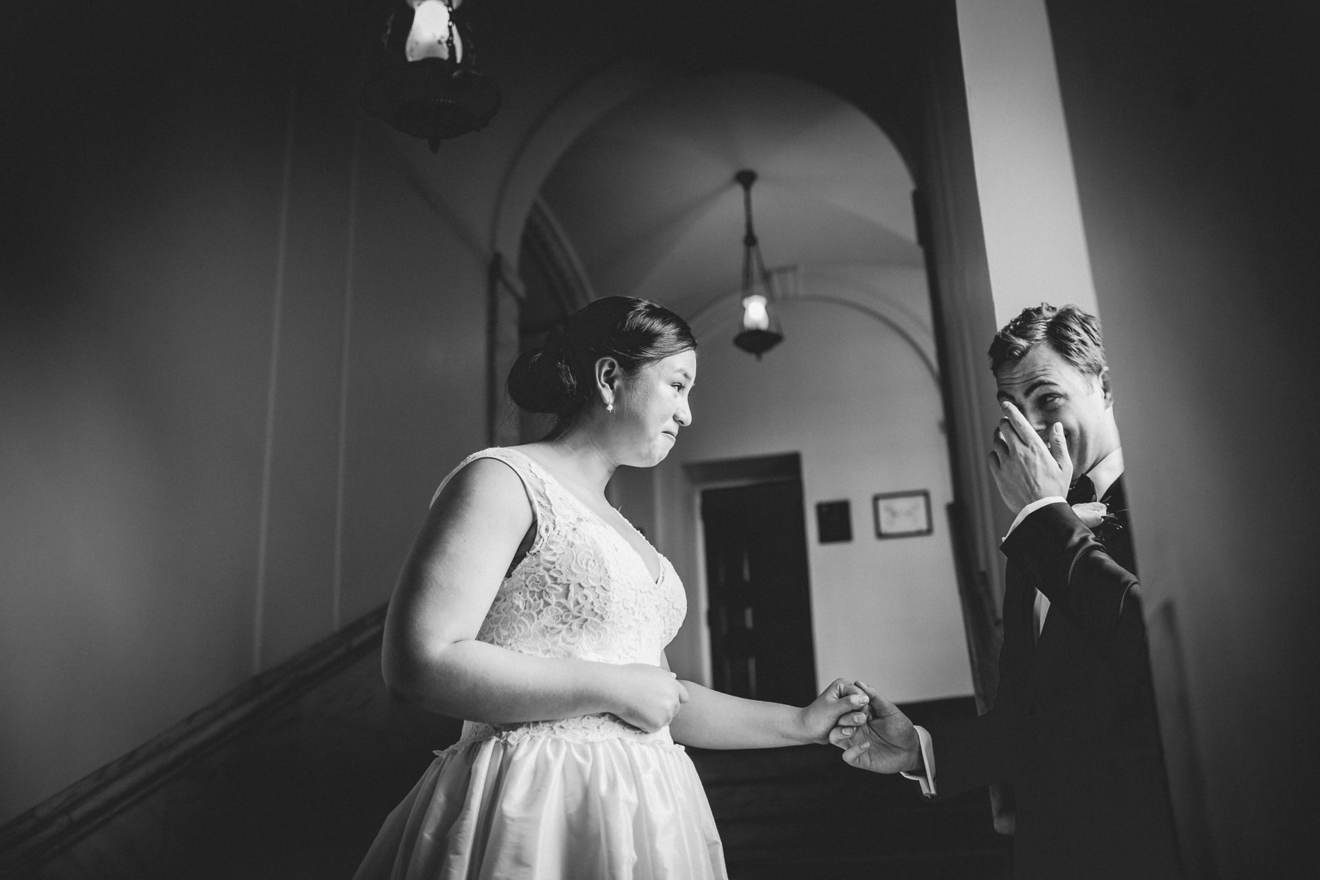 A bride and groom holding hands with emotional looks of love on their faces in a sidelit shadowy hallway. The groom wipes tears from his eyes.