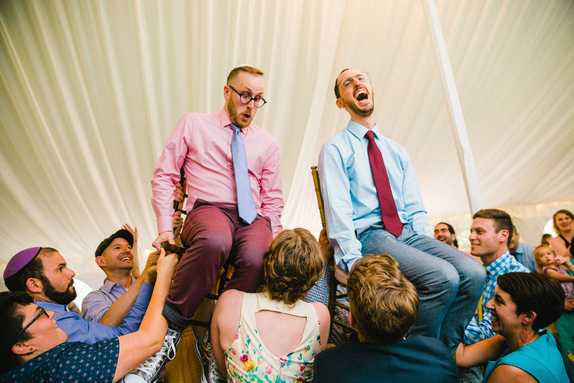 Two grooms raised overhead in chairs for the chair dance in a party tent with looks of shock and joy on their faces