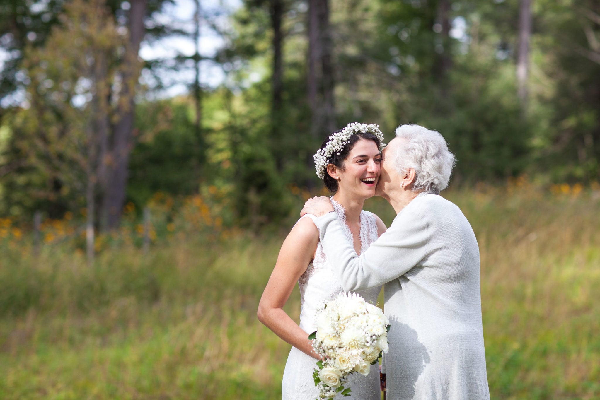 a bride with a white flower crown being kissed by a grandmother in a meadow surrounded by trees