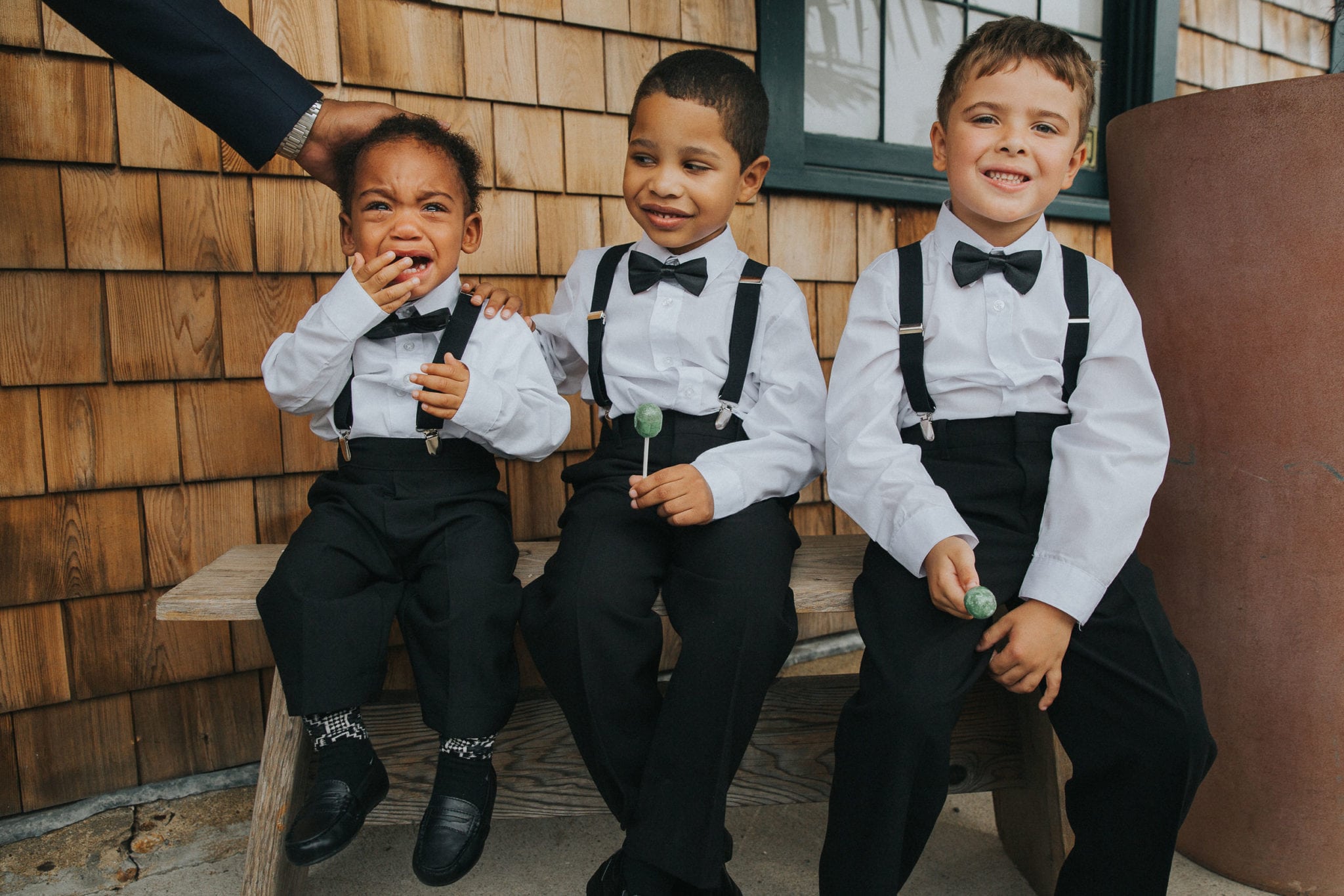 three little kids in white shirts, black pants, suspenders, and bow ties sitting on a bench near a wood shingled wall. The smallest one is crying while the middle one tries to comfort.