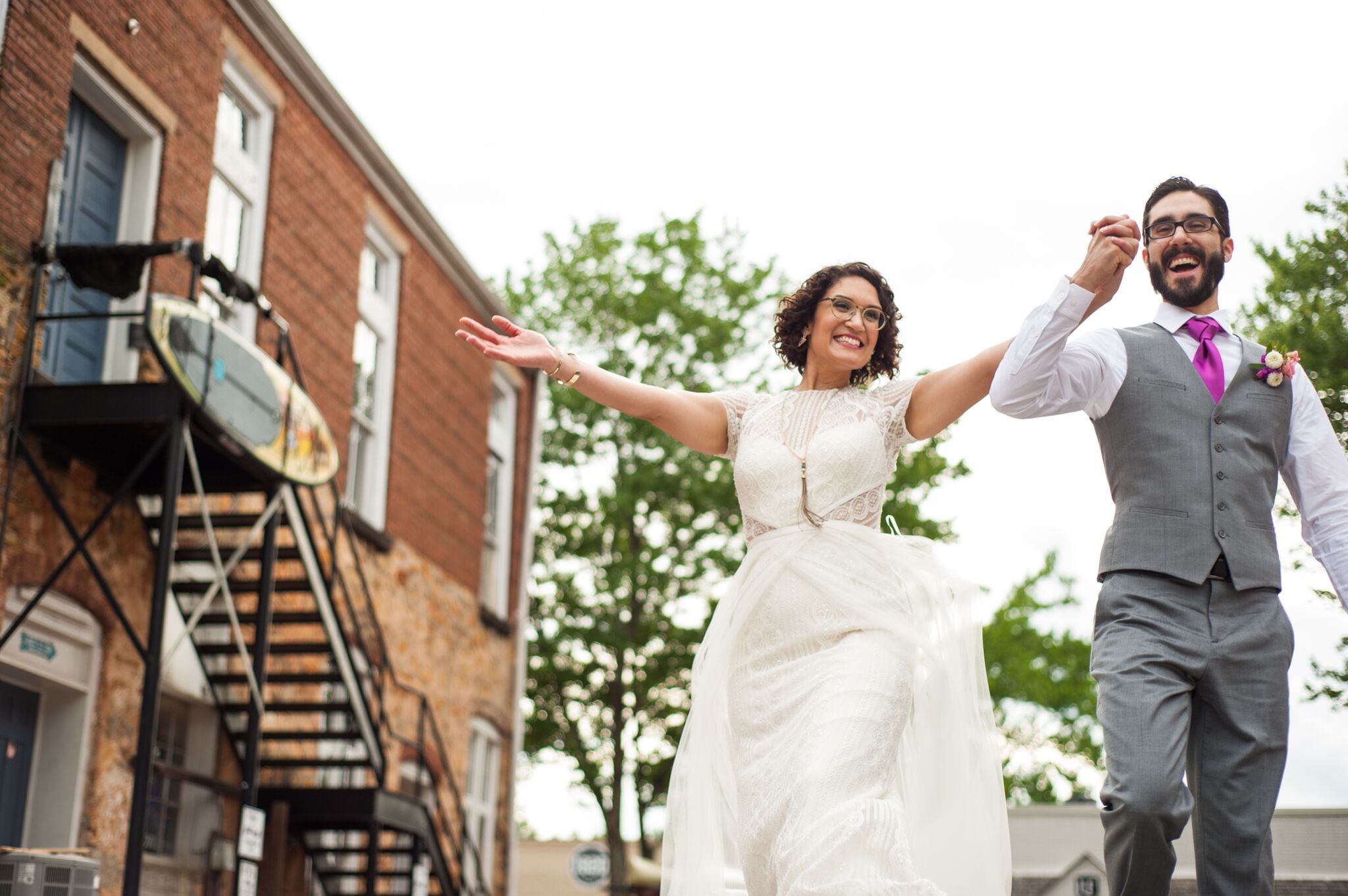 A couple in wedding clothes hold hands and raise them victoriously as they happily parade down the street