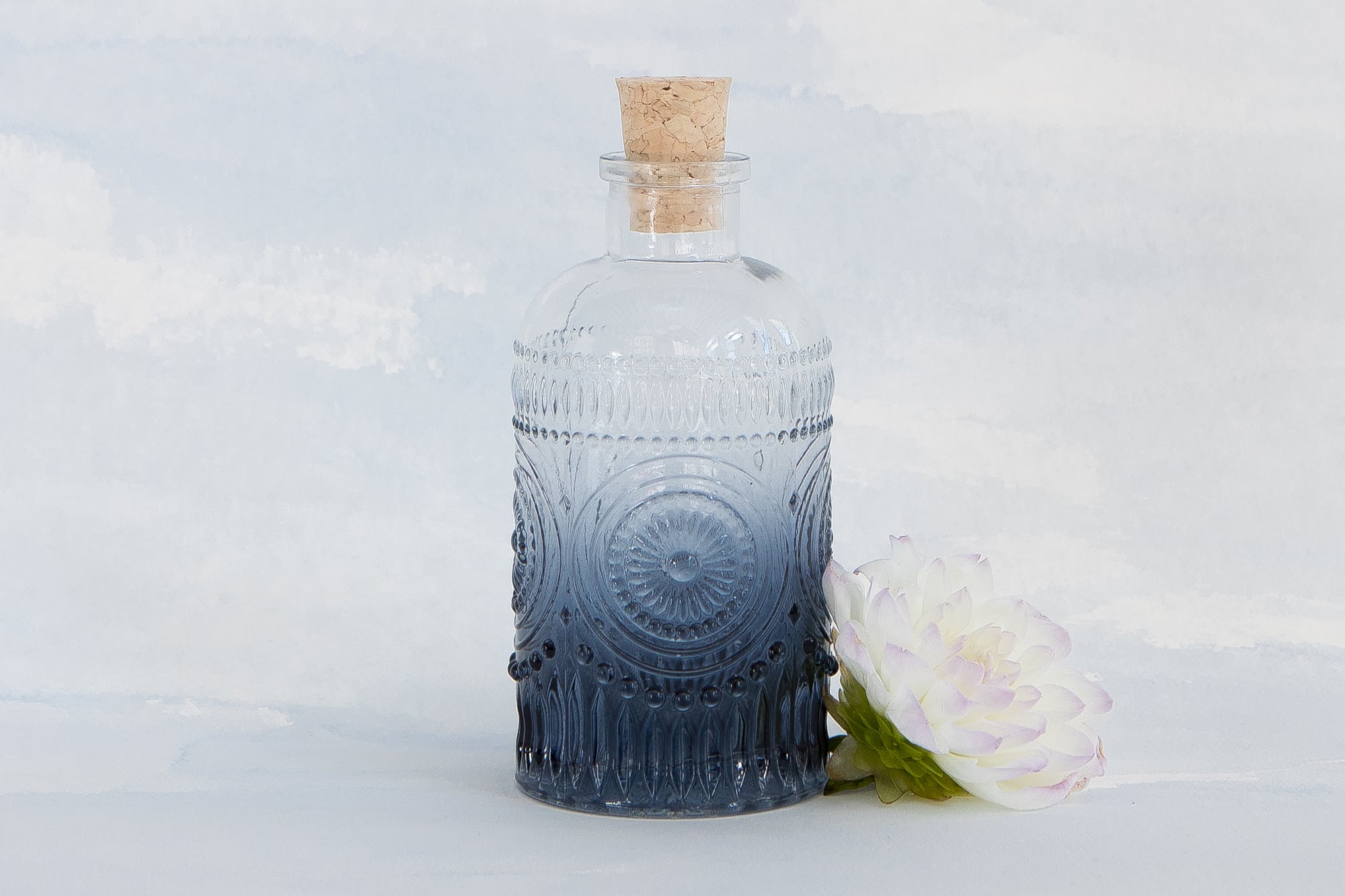 a navy blue bohemian inspired glass keepsake bottle styled on a blue watercolor backdrop with a single flower at the foot of the bottle