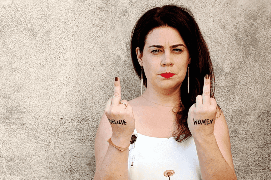 A brunette woman in a white floral print sundress stands in front of a dirty stucco wall with both middle fingers raised. Black marker on her hands reads, "Believe Women"