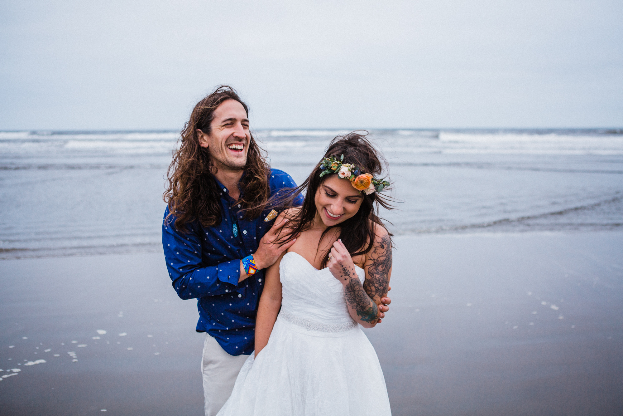 A man and a woman laugh together on the beach with waves crashing behind them