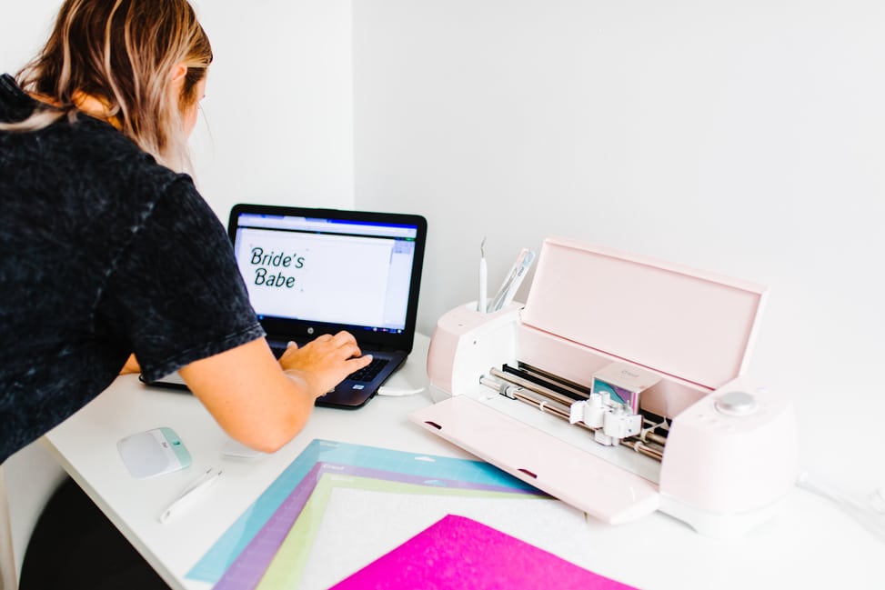 A woman using a laptop to design text to cut next to her Cricut Explore Air 2 Machine from Michael's Weddings