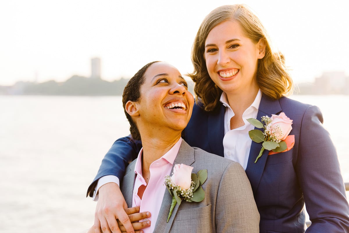 Kelly Prizel photo of two smiling women standing on a waterfront, one has her arm around the other in front who looks back at her. Both are wearing suits with rose boutonnieres, the one in front is grey with a pink shirt, the one behind is navy with a white shirt.