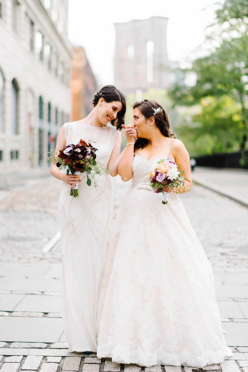 Kelly Prizel photo of two brides standing on a cobblestone street with the Brooklyn Bridge in the distance. They are both wearing wedding dresses and holding small bouquets and one is kissing the hand of the other.