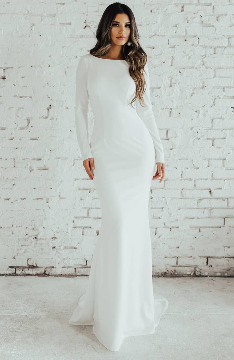brunette woman wearing a long sleeve wedding gown standing in from of a white brick wall