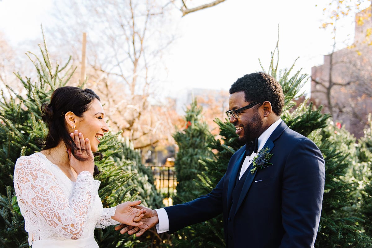 Kelly Prizel photo of black groom with glasses and a navy blue tuxedo reaches his hand out to bride's hand. She is wearing a white lace long sleeved wedding dress and has her other hand to her face as her brown hair is blowing in the wind.