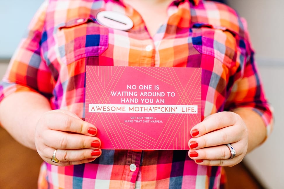 A woman in a brightly colored plaid shirt with red painted fingernails holds a pink card that reads: "No one is waiting around to hand you an awesome motha'f*ckin' life. Get out there + make that shit happen."