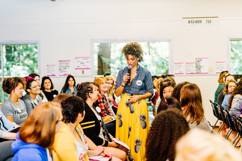 Cyndie Spiegel wearing a yellow pinapple print skirt and chambray button down top holds a microphone and stands in the audience during her keynote speech at The Compact Camp