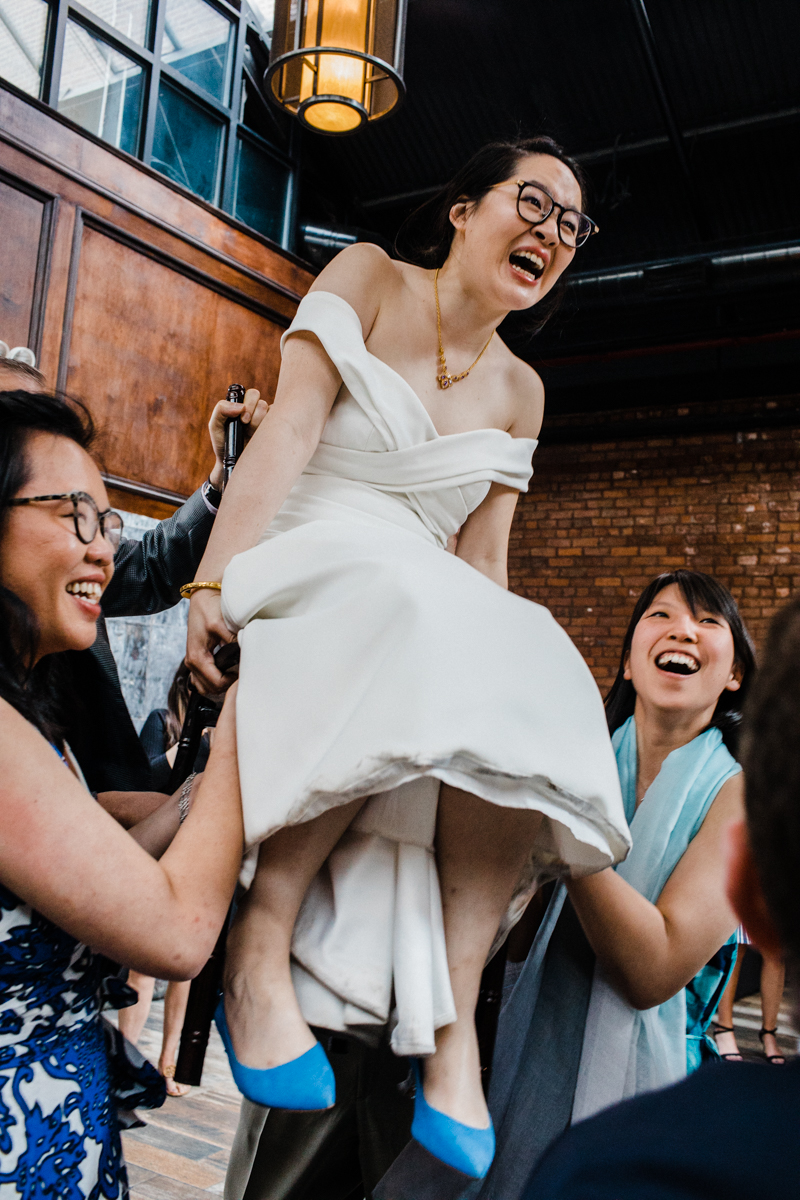 Bride with glasses and blue shoes is laughing as she is lifted into the air on a chair