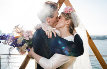 A Corey Torpie photo of two people in wedding kissing in a tight embrace in front of a wooden triangular altar piece, overlooking the water.