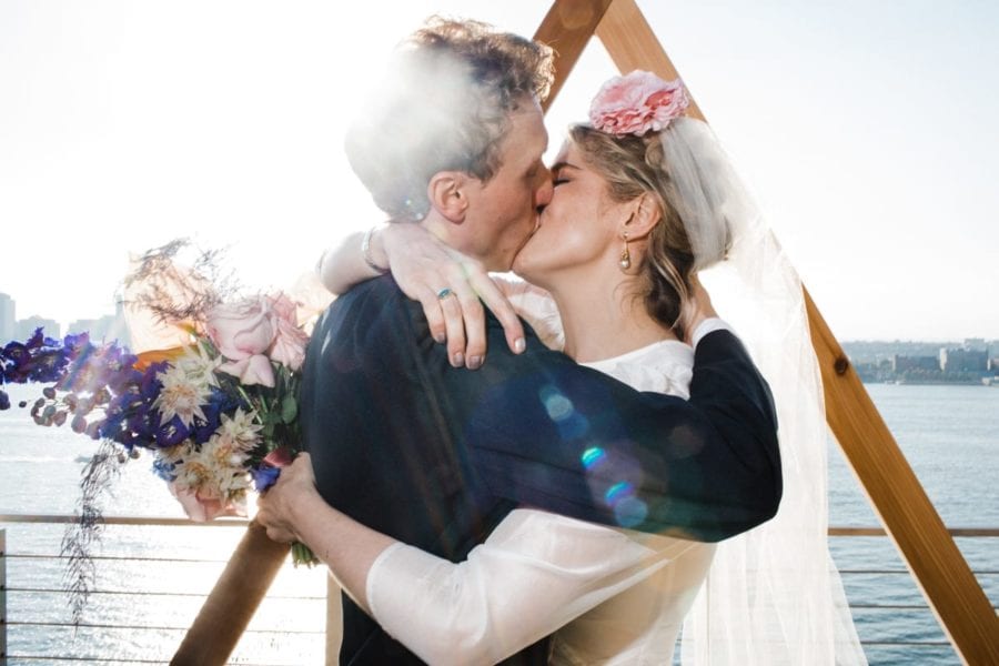 A Corey Torpie photo of two people in wedding kissing in a tight embrace in front of a wooden triangular altar piece, overlooking the water.