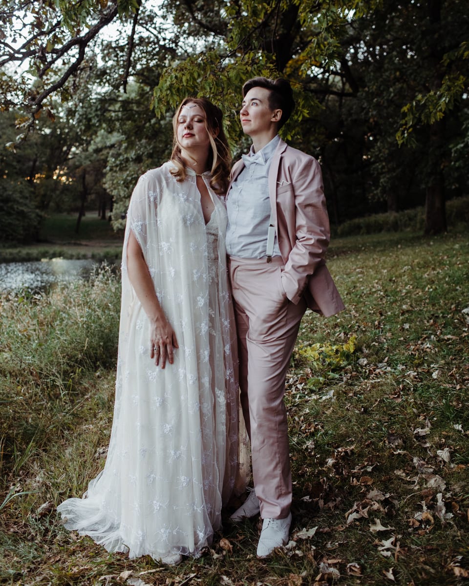 Corey Torpie photo of a couple standing in a forest with their arms around each other. One is wearing a lace caftan and the other a pink suit.
