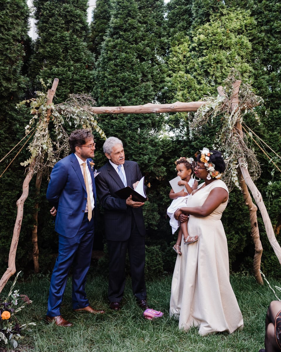 couple standing under a chuppah with their officiant. The bride is holding a child who's also wearing a flower crown.