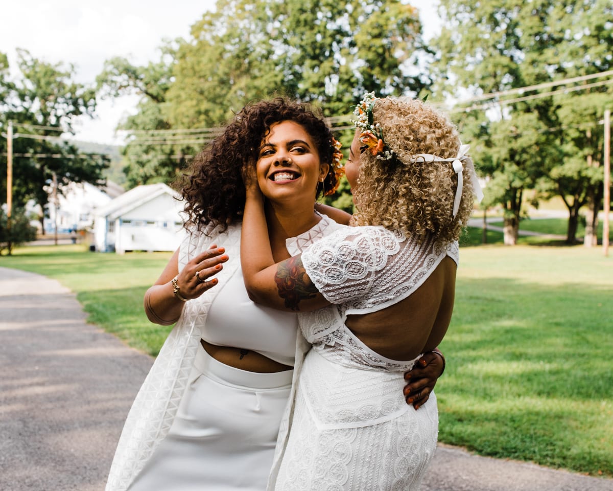 two brides standing on a lawn hugging and smiling, one has a flower crown and the other is wearing a crop top wedding dress