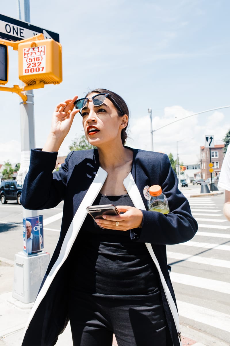 Corey Torpie photo of Alexandria Ocasio-Cortez crossing a street while lifting her sunglasses with one hand, phone in other hand