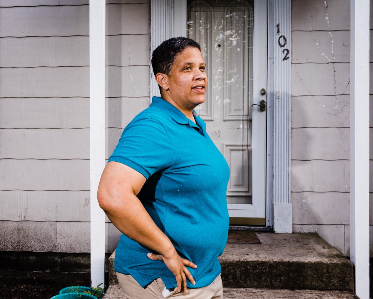 Corey Torpie photo of Kerri Evelyn Harris in a blue polo shirt, standing in front of a door to a house with white siding