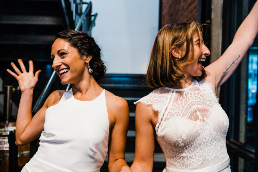 A Corey Torpie photo of two women in white wedding dresses holding hands, smiling, and waving in opposite directions