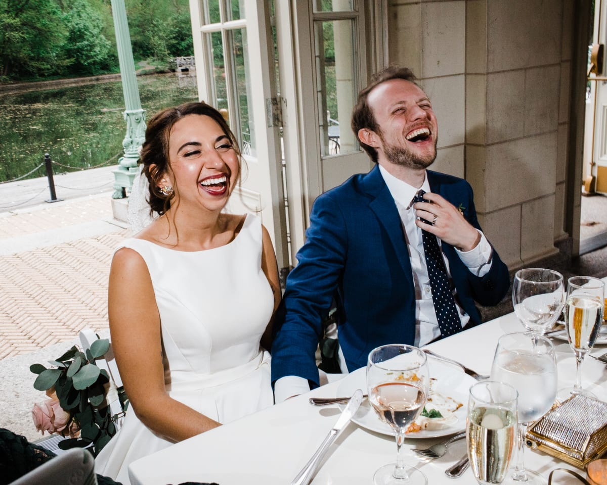 Bride and groom sitting at table at Brooklyn Boat House, holding hands and laughing