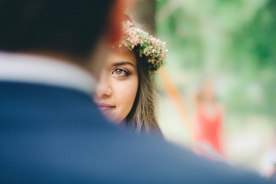 A woman with a flower crown, hazel eyes, and brown hair looks forward toward you at the man she is marrying who is out of focus with his back to you, wearing a blue skirt.